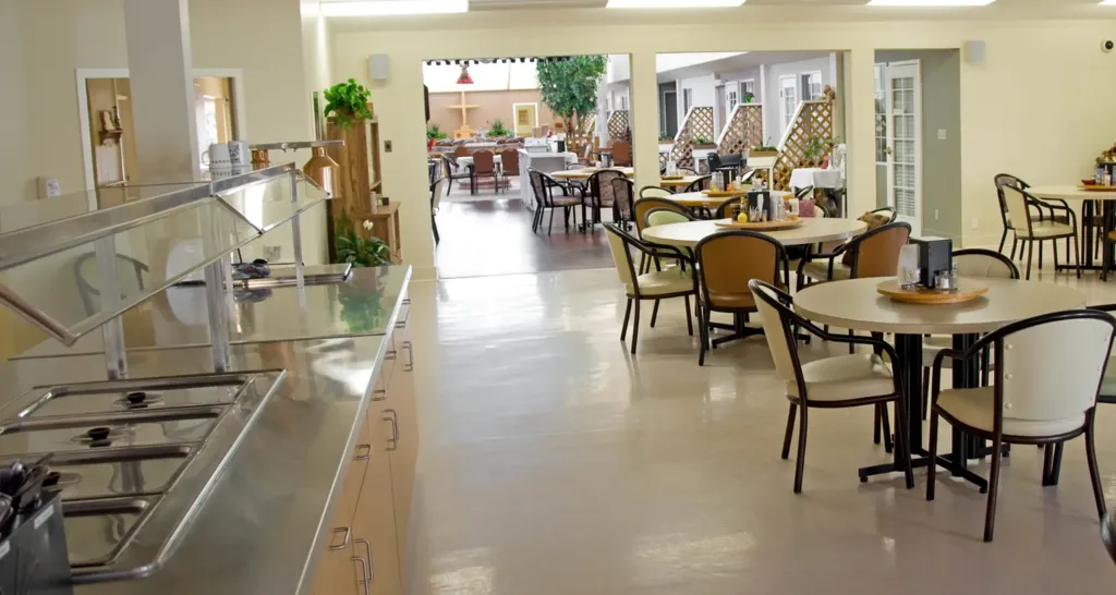 Bright senior living community dining room with cafeteria-style serving line, stainless steel food stations, round dining tables, and spacious seating area prepared for resident meal service.