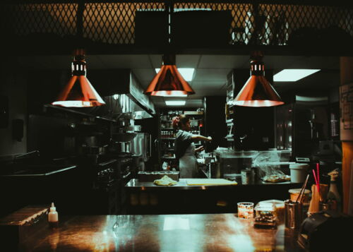 Chef working in a dimly lit commercial kitchen, surrounded by cooking equipment and supplies, with warm lights overhead.