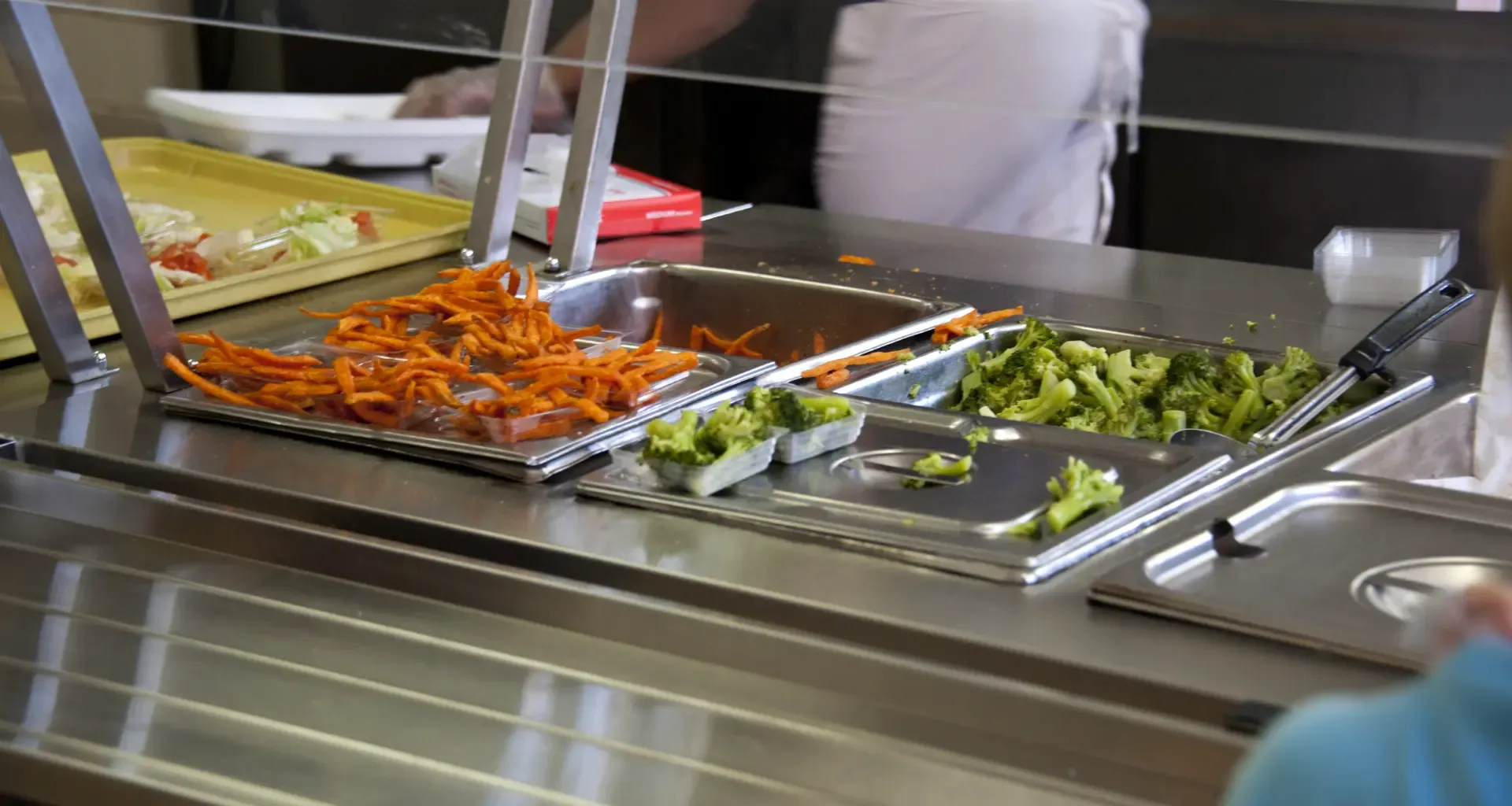 School cafeteria serving line with stainless steel steam table holding cooked carrots and broccoli, partially filled food pans, and cafeteria staff preparing meals during lunch service.
