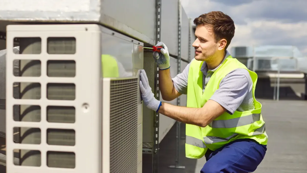 HVAC technician in high-visibility vest repairing rooftop commercial AC unit under sunny sky