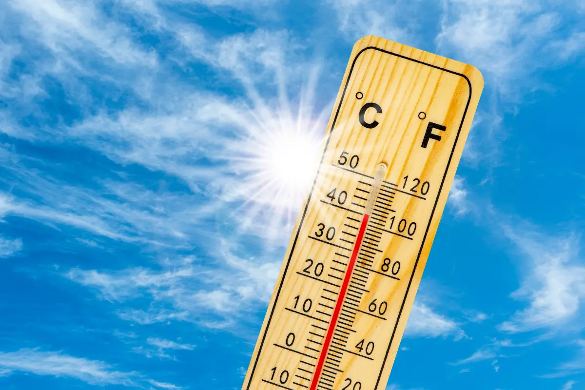 Wooden thermometer showing high temperatures in both Celsius and Fahrenheit, angled against a bright blue sky with the sun shining intensely—symbolizing extreme summer heat or a heatwave.